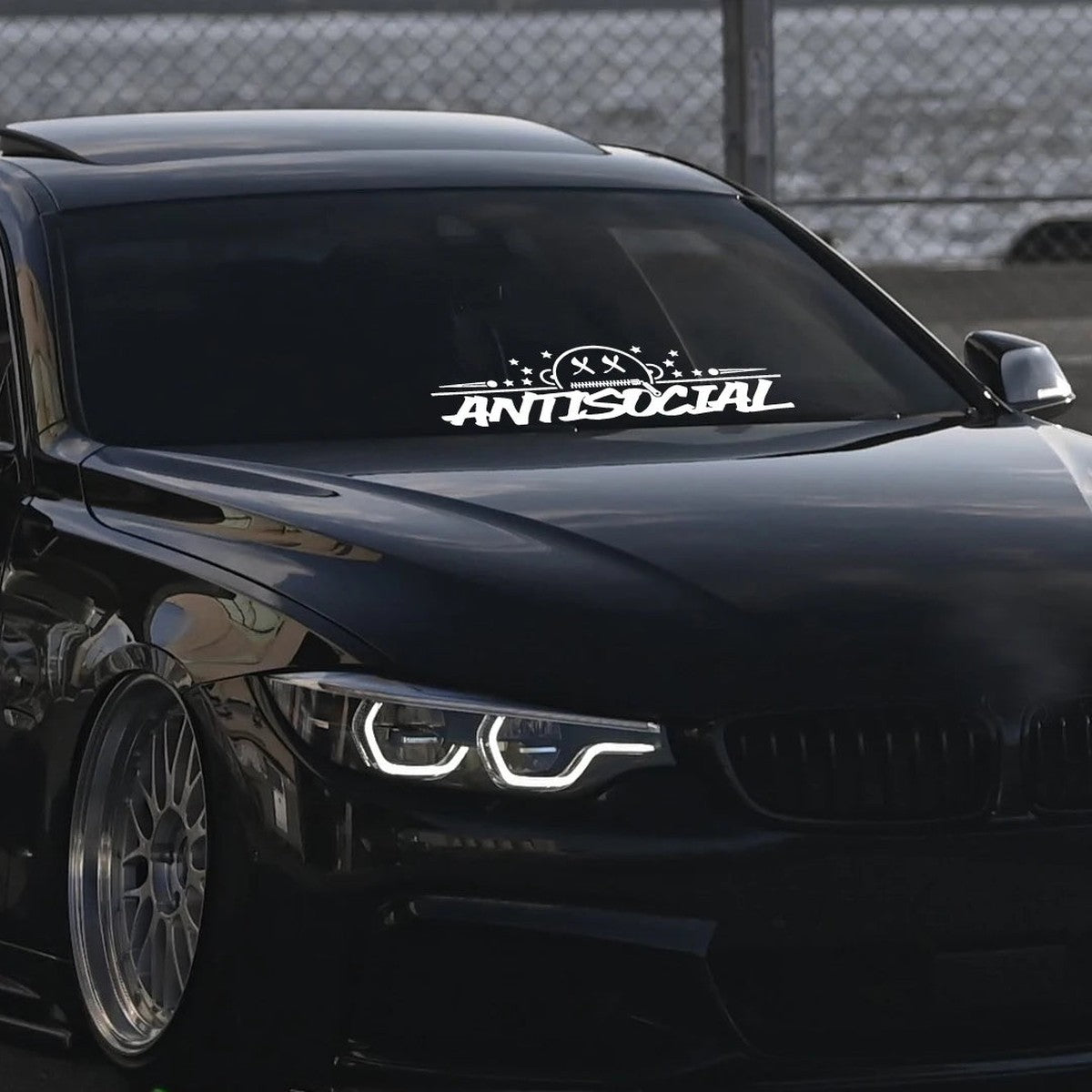 Antisocial windshield banner decal on a black BMW with angel-eye headlights