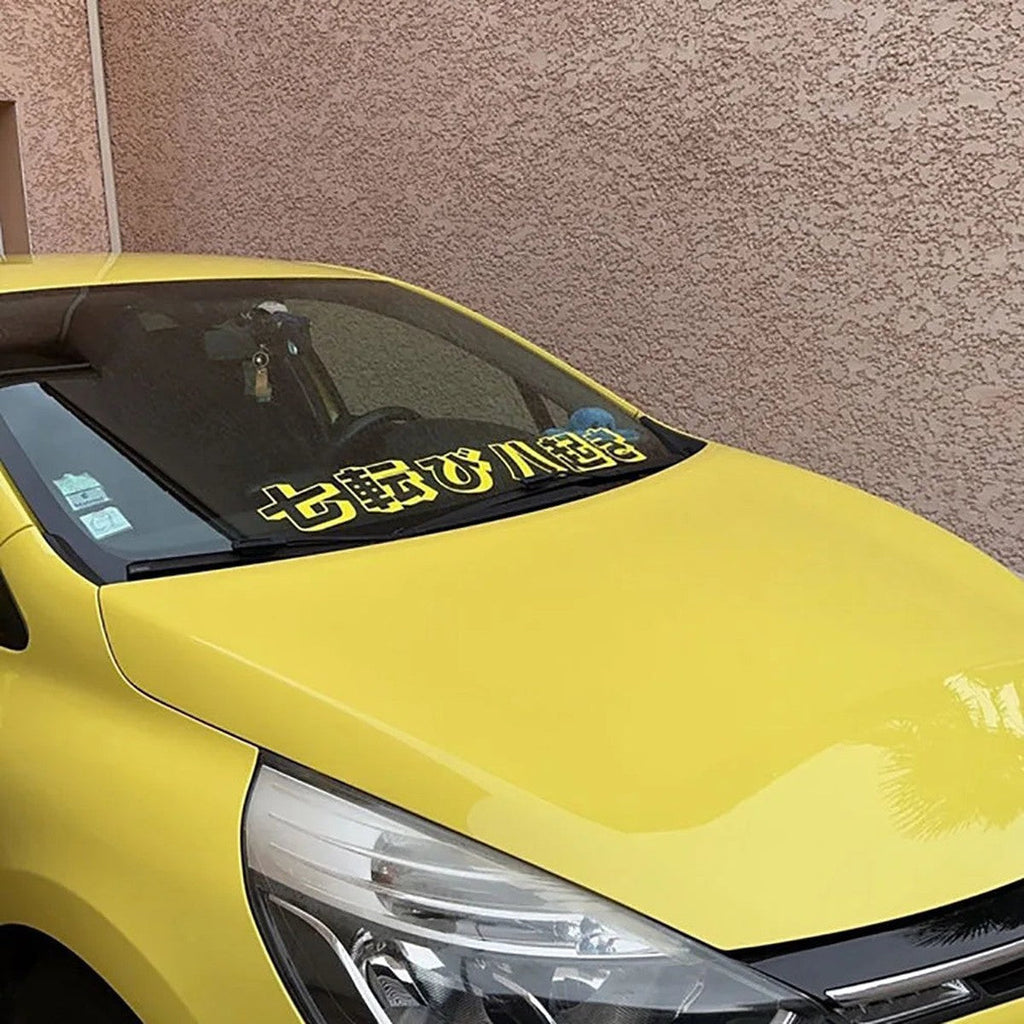 Yellow kanji windshield banner decal on a yellow hatchback, front three-quarter view